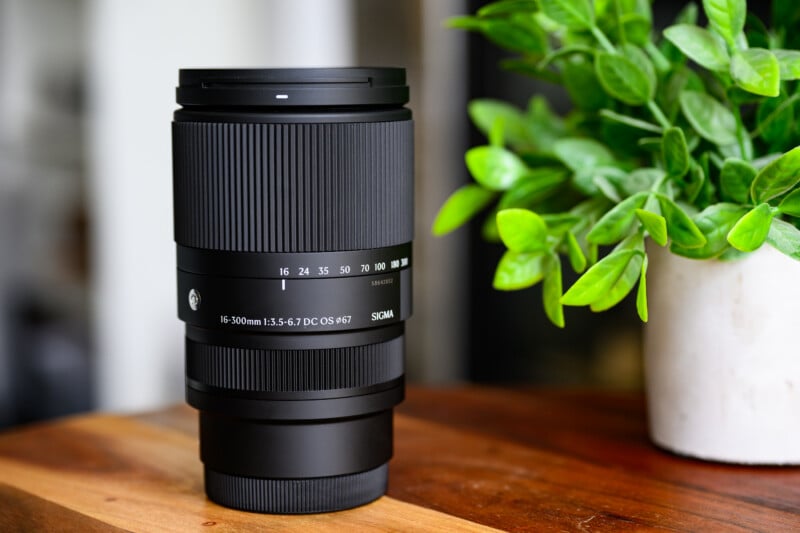 A black Sigma 16-300mm camera lens sits on a wooden surface. The lens displays focal length markings and brand details. A potted plant with green leaves is blurred in the background, adding a touch of color.
