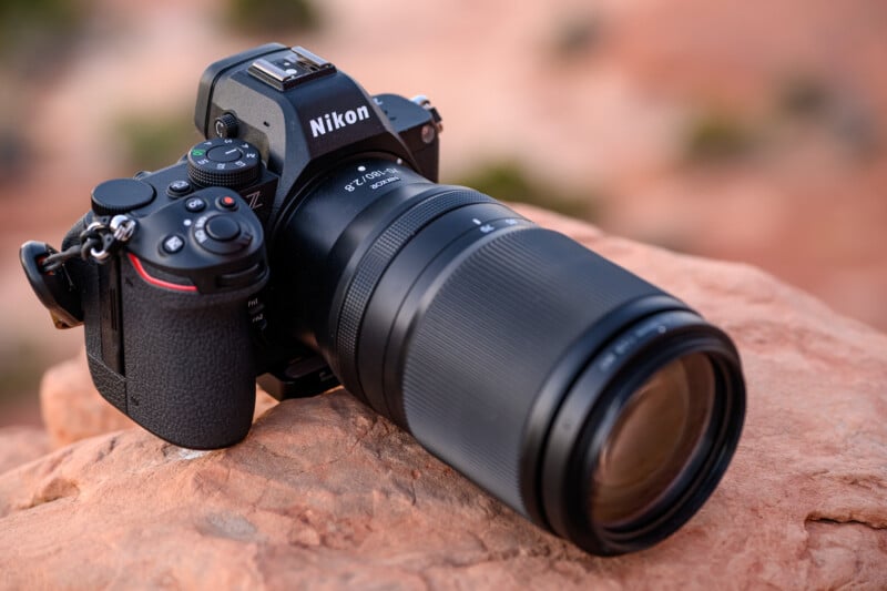 Close-up of a Nikon Z camera with a large telephoto lens positioned on a rocky, reddish surface. The camera is angled slightly to the left, with the ambient light suggesting an outdoor setting, possibly during sunset.