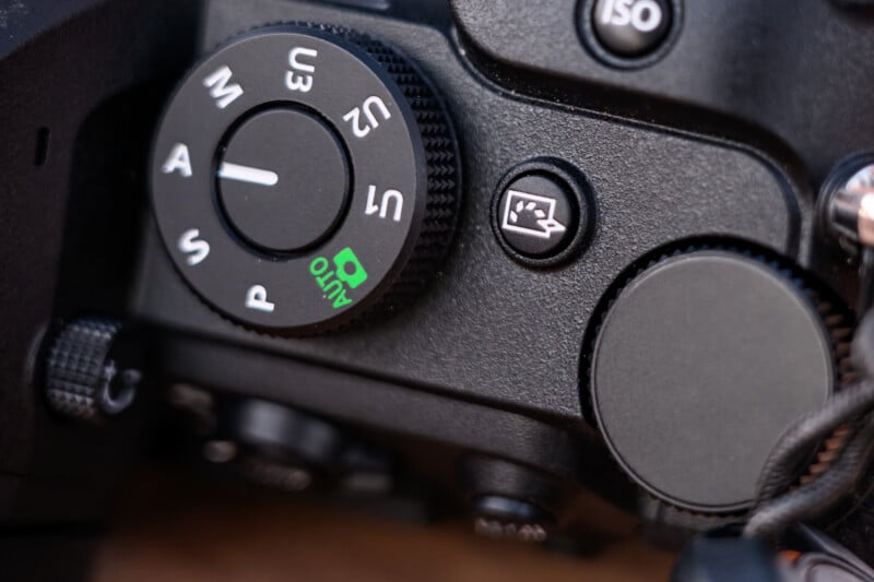 Close-up of a camera mode dial showing settings like M (Manual), A (Aperture Priority), S (Shutter Priority), P (Program), and Auto. The dial is next to other controls in the corner of a black digital camera.