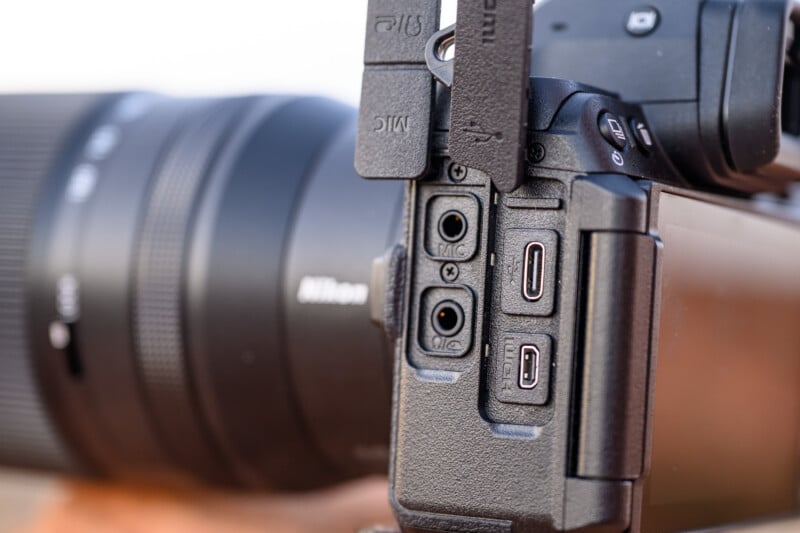 Close-up of a Nikon camera showing side ports, including two audio jacks, two USB-C ports, and part of the camera's screen. The black lens with the Nikon logo is visible in the background.