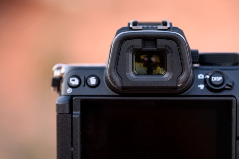 Close-up of the back of a digital camera focusing on the viewfinder and control buttons. The camera's LCD screen is off. The background is softly blurred in warm tones.