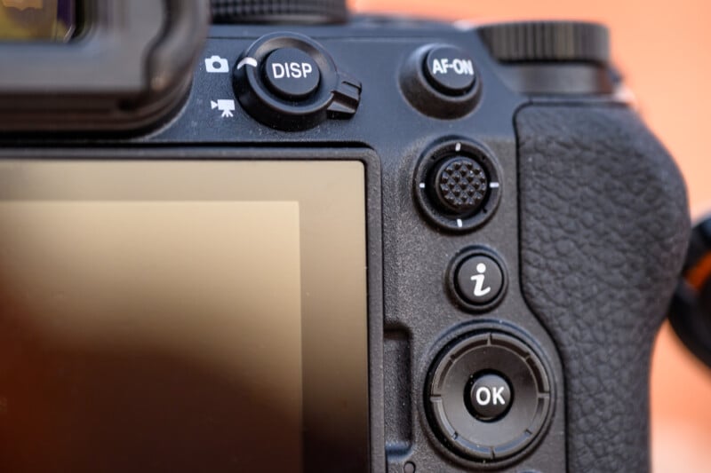 Close-up of a camera's rear controls including DISP, AF-ON, and OK buttons. Textured grip is visible on the right with a screen partially in view on the left, all set against a blurred background.