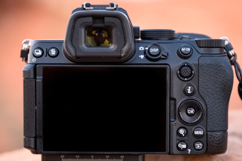 A close-up of the back of a digital camera displaying various buttons, a control dial, and a screen. Features include a viewfinder, menu, and info buttons, and a textured grip on the right side. Background is softly blurred.