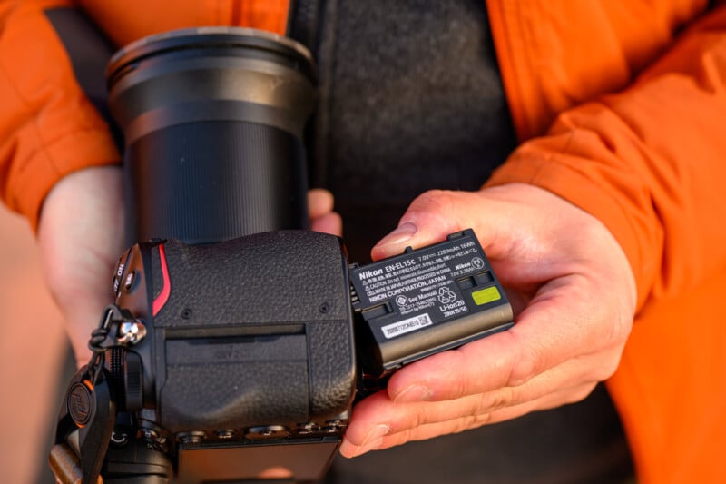 Person in an orange jacket holds a Nikon camera with one hand while inserting a Nikon EN-EL15c battery with the other. The focus is on the battery label showing specifications.