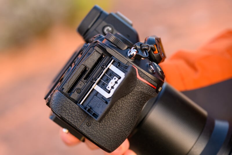 Close-up of a person holding a DSLR camera with an exposed memory card slot. The camera has a textured black grip and a red accent line. The background is blurred, suggesting an outdoor setting.