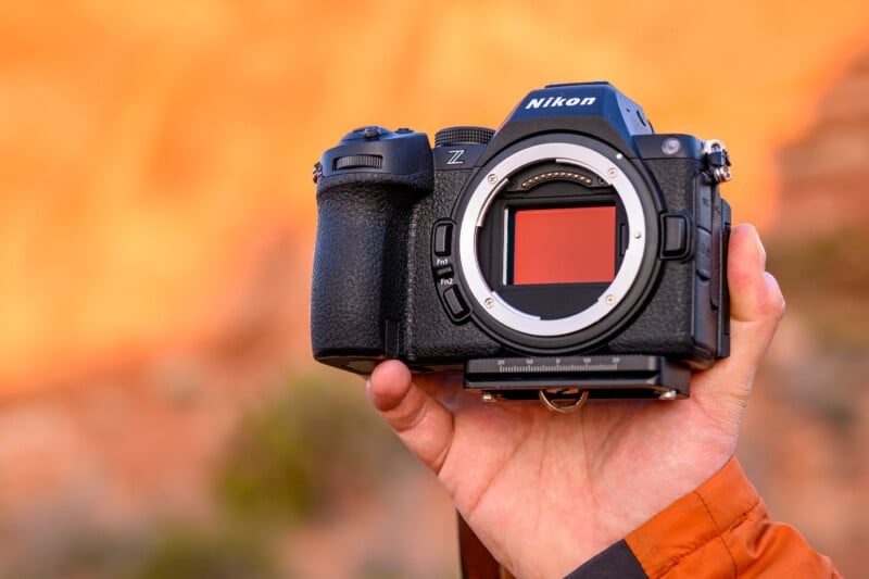 A hand holds up a Nikon Z series mirrorless camera against a blurred outdoor background with warm, orange hues. The lens is removed, revealing the camera's sensor. The person wears an orange jacket.