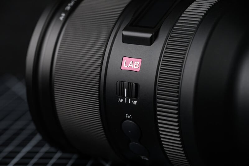Close-up of a camera lens with a pink "LAB" sticker. It features a switch labeled "AF" for autofocus and "MF" for manual focus, and buttons labeled "Fn1" and "TP" below. The lens is resting on a grid-patterned surface.