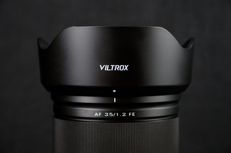 Close-up of a Viltrox camera lens with "AF 35/1.2 FE" markings. The lens features a black finish and a petal-shaped lens hood, set against a dark background.