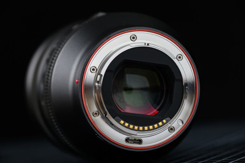 Close-up of a black camera lens with a metallic mount, featuring a red ring and electronic contacts. The lens is placed on a dark surface with a soft-focus background.