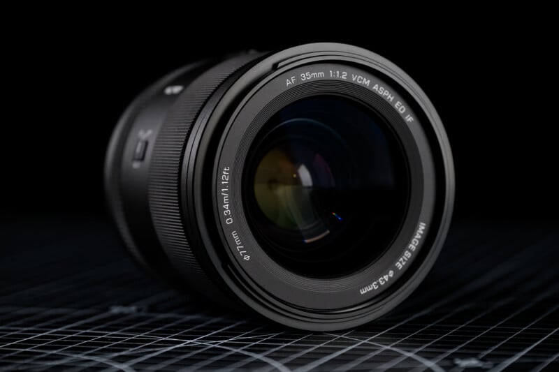 Close-up of a camera lens resting on a grid-patterned surface. The lens has clear markings indicating it is a 35mm with specifications like 1:1.2, VCM, and others. The background is dark, emphasizing the lens's intricate design.