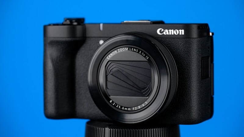 A black Canon digital camera with a closed lens cover is positioned on a tripod against a bright blue background.