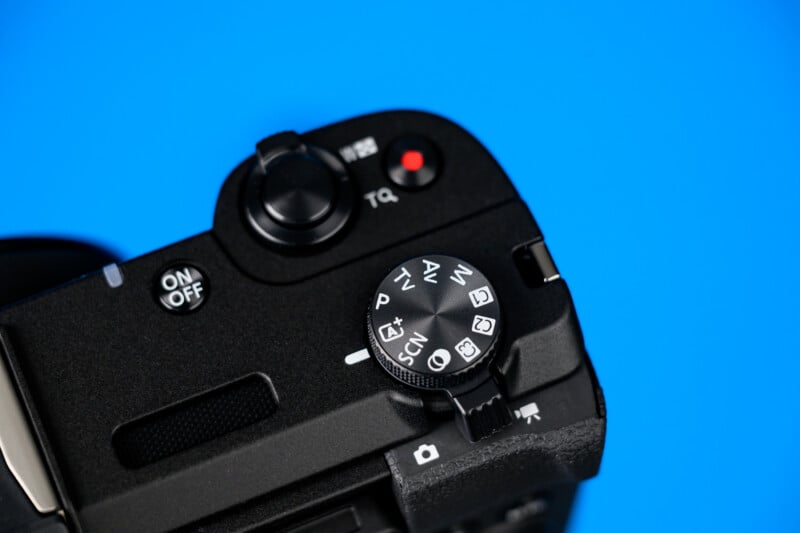 Close-up of the top of a black digital camera against a blue background, showing the mode dial with various settings, the on/off switch, and a red video record button.