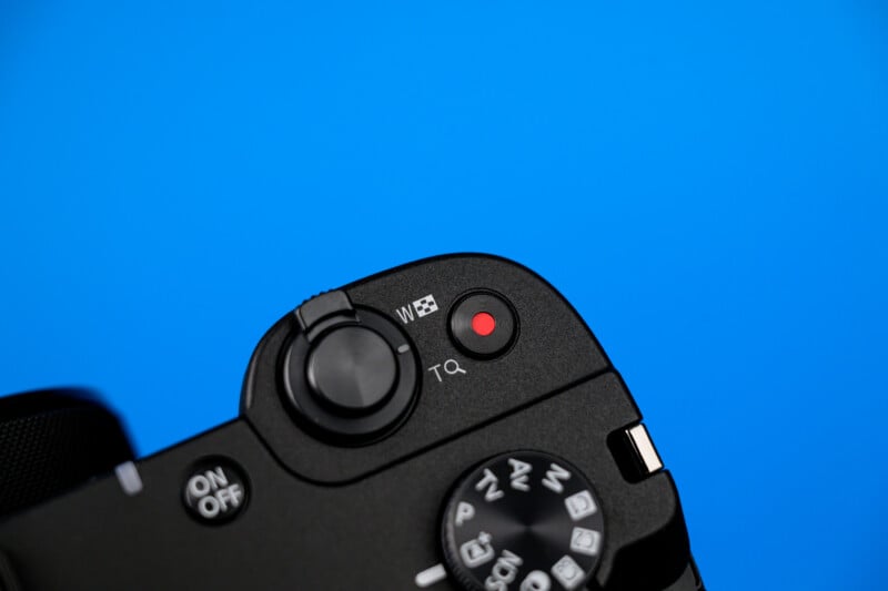 Close-up of the top of a black digital camera with a red record button, mode dial, and on/off switch, set against a bright blue background.