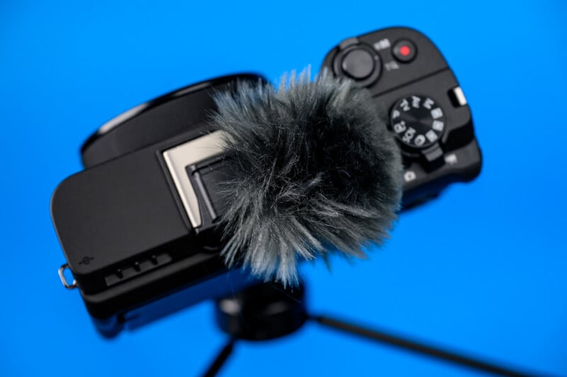 A black digital camera with a furry windscreen attached to its microphone is mounted on a tripod against a solid blue background.