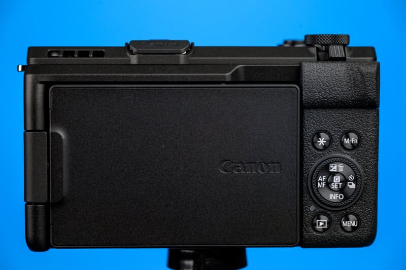The back view of a black Canon digital camera with the screen closed, various control buttons visible, and a blue background.