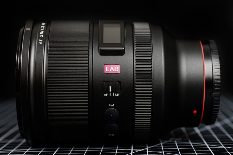 A black camera lens with "AF 35/1.4 Z" labeled on it, featuring a small screen and red ring. It rests on a grid-patterned surface, with the label "LAB" visible near the middle.