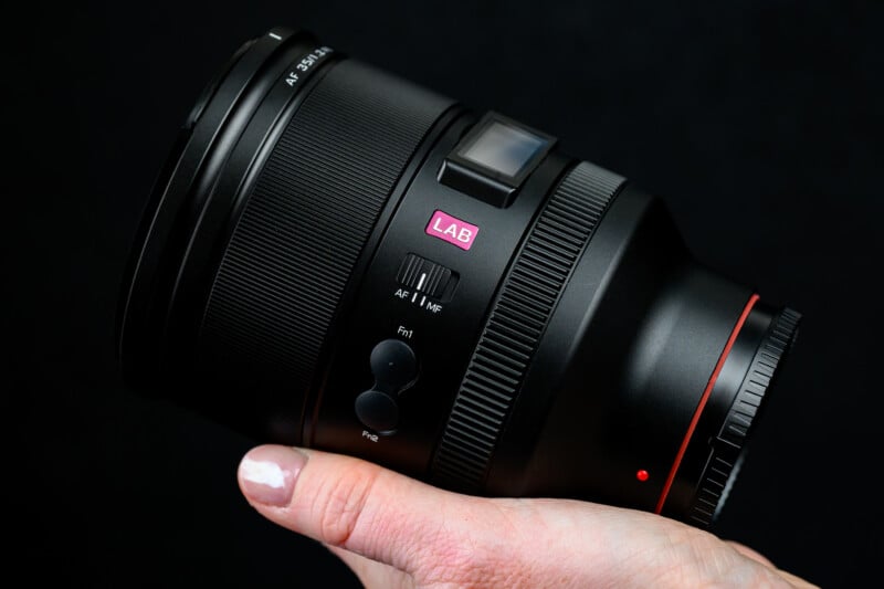 Close-up of a person holding a black camera lens with their hand. The lens features various controls and markings, including a pink "LAB" sticker. The background is solid black.