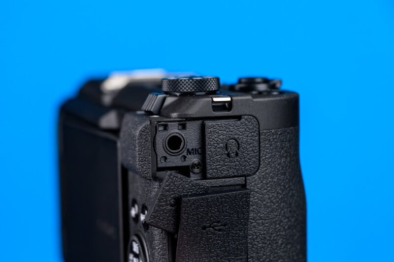 Close-up of the side of a black digital camera against a blue background, showing open ports labeled "MIC" and a headphone icon, with textured grip and control dials visible.