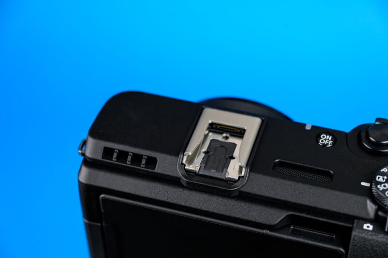 Close-up of a black digital camera against a blue background, showing the top part with a metal hot shoe mount, an on/off switch, and control dials.