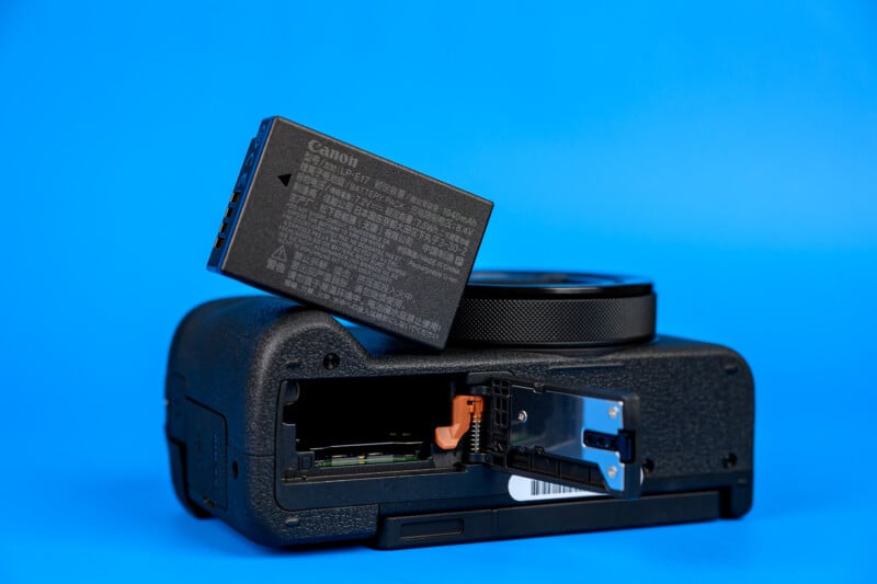A Canon camera body with its battery compartment open, displaying a rechargeable Canon battery above the compartment, set against a plain blue background.