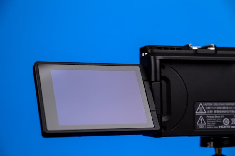 A digital camera with its flip-out LCD screen extended, facing left against a solid blue background. The screen is blank and the camera's side panel with labels is partially visible.