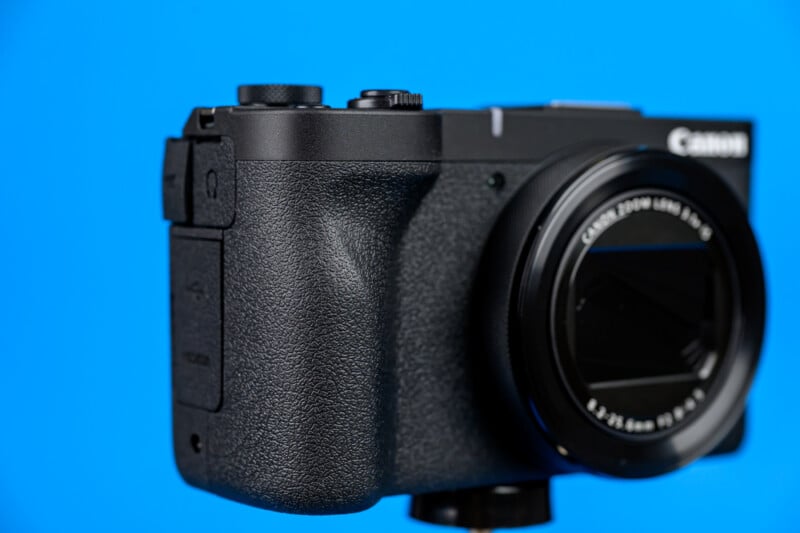 A close-up of a black Canon digital camera against a solid blue background, showing the textured grip, lens, and part of the camera body.