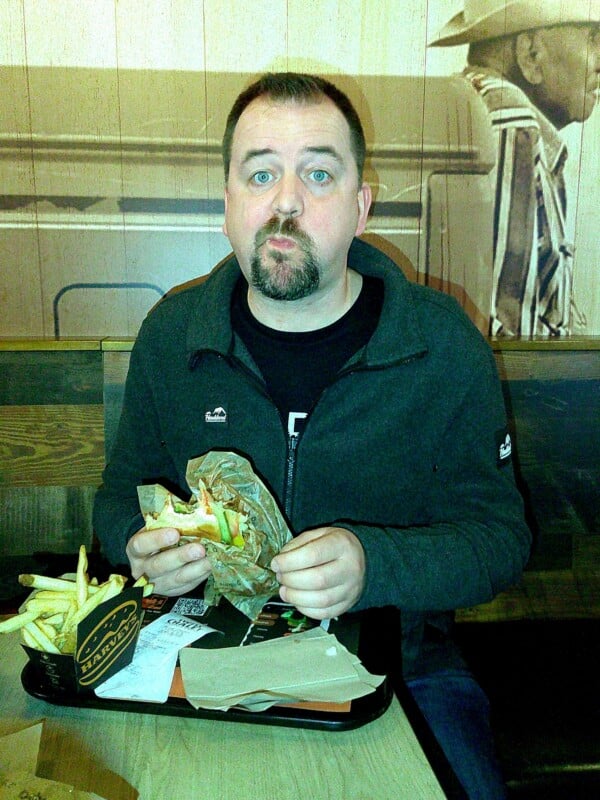 A man with a surprised expression sits at a table in a restaurant, holding a sandwich. There's a tray with fries and a drink in front of him. The background features wall art of a person in a hat.