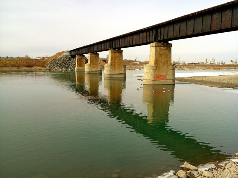 A rusted metal railroad bridge spans across a calm river with clear reflections. The bridge is supported by concrete pillars. The riverbank is rocky, and the sky is overcast. Industrial buildings can be seen faintly in the background.