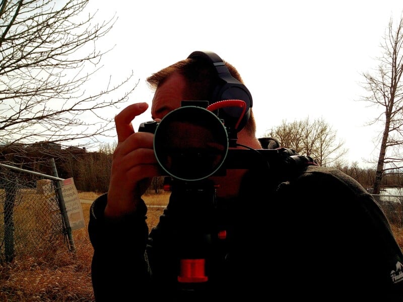 Person wearing headphones and holding a camera on a tripod outdoors. Bare trees and a fence are visible in the background against a bright sky.