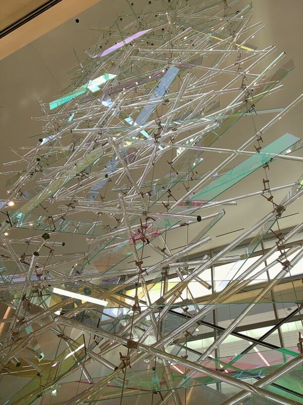 A large, intricate sculpture made of metallic rods and transparent panels in various colors stands inside a modern building. The artwork extends upwards, creating a dynamic, geometric pattern that interacts with the ambient light.