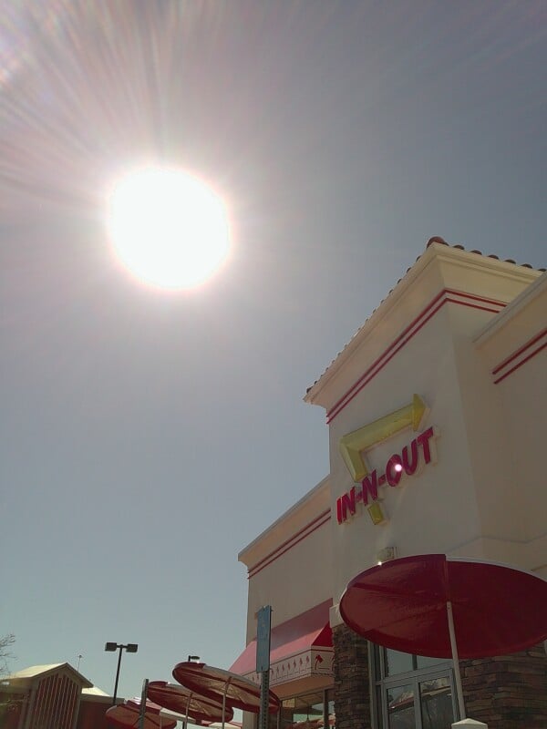 A bright sun shines in a clear sky above an In-N-Out Burger restaurant. The building features the chain's red and yellow sign, with red patio umbrellas visible below.