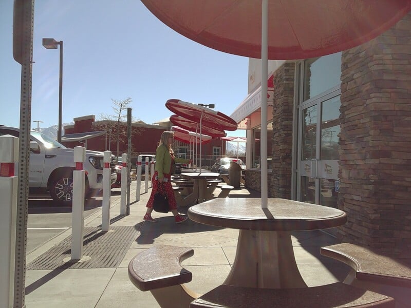 A person walks past outdoor picnic tables with red umbrellas at a restaurant. The sky is clear and sunny, and cars are parked nearby. The building has stone accents and large windows.