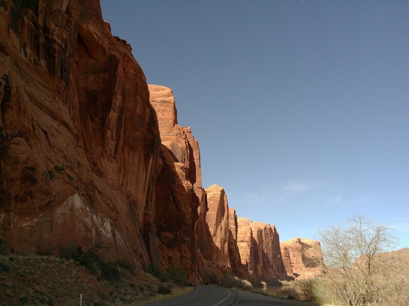 A scenic view of large red rock cliffs under a clear blue sky. A road winds along the base of the cliffs, with sparse vegetation and a few barren trees along the side.