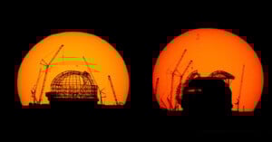 Two large telescope domes under construction are silhouetted against a bright orange sunset, with several cranes visible around the structures.