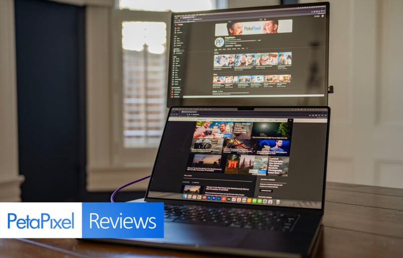 Dual-screen laptop on a table displays the Petapixel website. The setup includes a larger screen above the main laptop screen. The room has white walls and a partially open window with blinds.