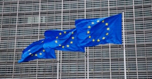 Three European Union flags with yellow stars on blue backgrounds fly in front of a modern building with a grid-like facade.