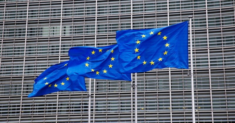 Three European Union flags with yellow stars on blue backgrounds fly in front of a modern building with a grid-like facade.