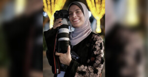 A smiling woman wearing a floral-patterned abaya and light-colored hijab holds a professional camera with a large lens. Warm, decorative lights glow in the blurred background at night.