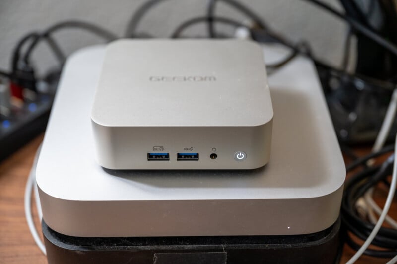 A silver mini PC sits on top of a larger, similar device. The mini PC has two USB ports, an HDMI port, a headphone jack, and a power button on its front panel. Cables are visible in the background.