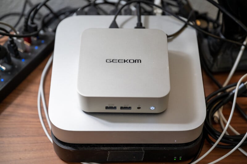 A compact silver device labeled "GEEKOM" sits on top of another similar-looking device. Both are surrounded by various cables on a wooden surface. The front of the GEEKOM device features two USB ports and a small power light.