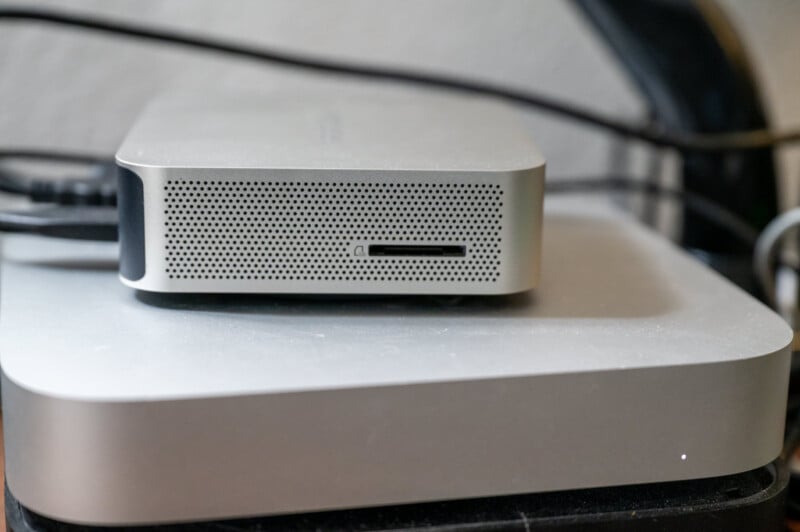 A compact, silver external hard drive with ventilation holes on the front rests on a slightly larger, matching metallic device. Cables are visible in the background, suggesting connectivity or power.