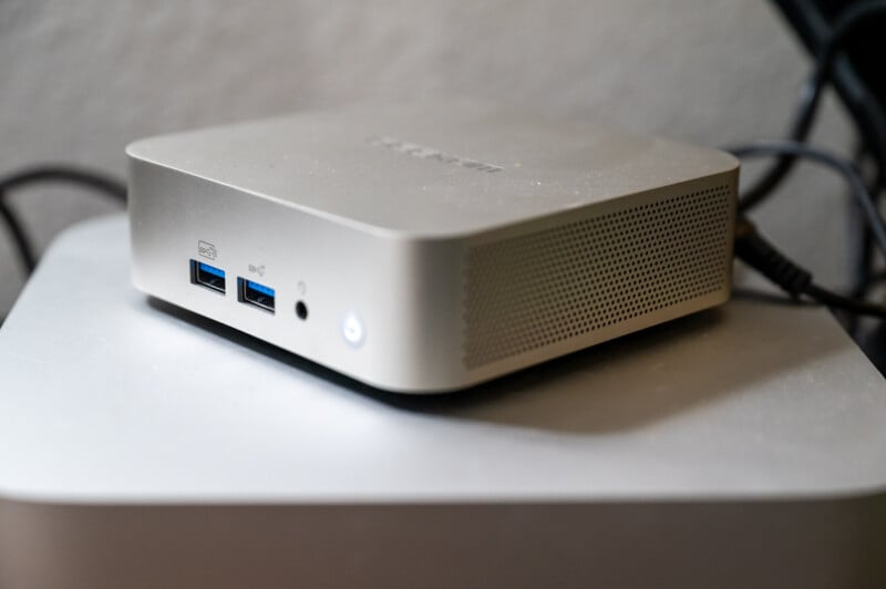A compact grey computer sits on a surface. It features USB ports and a power button on the front. The design is sleek and minimalist, with perforations for ventilation on the side. Cables are connected at the back.