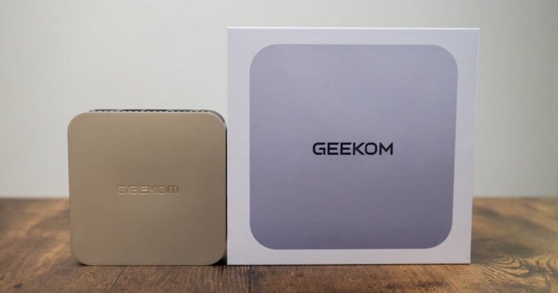 A small gold Geekom device sits on a wooden surface next to its white box, which has a gray square and the word "GEEKOM" on the front. The background is plain light gray.