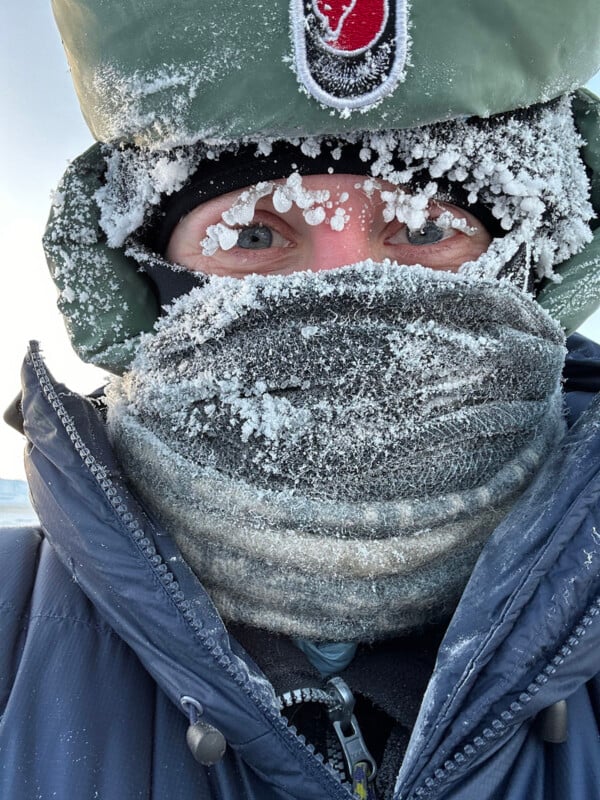 A person bundled in winter clothing with their face mostly covered in frost and snow, eyes visible between a woolen scarf and a hat with a red and white patch. Only the person's eyes are visible.