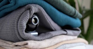 A small camera peeks out from inside a rolled-up gray sweater, which is stacked on top of other folded sweaters in green, blue, and beige tones. A leafy plant is blurred in the background.