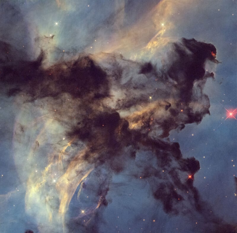 Dark clouds of interstellar dust and gas create dramatic shapes against a backdrop of stars and glowing nebulae in deep space, with some red and yellow highlights visible throughout the cosmic scene.