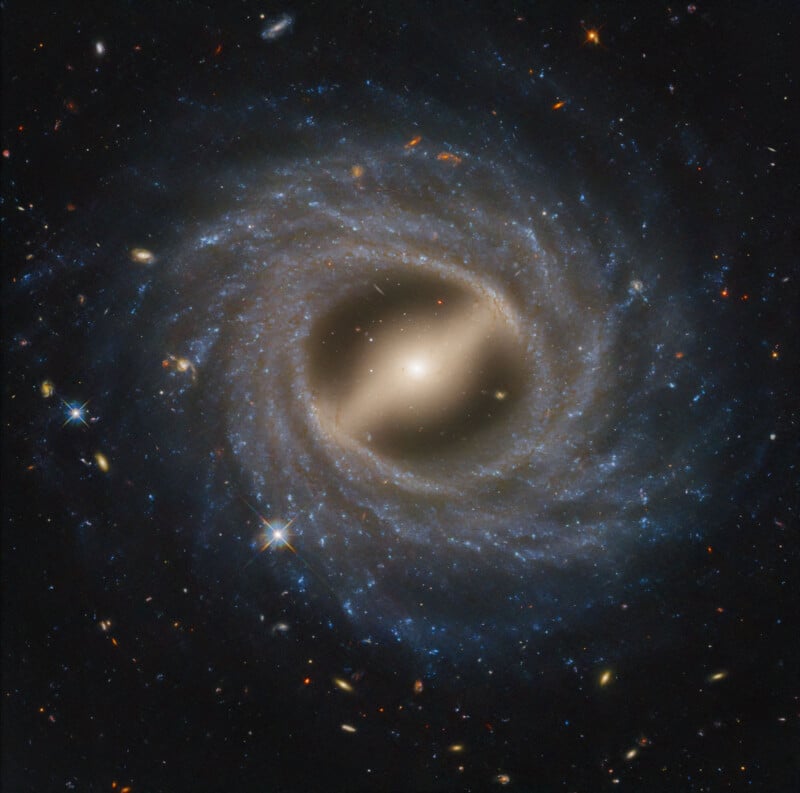 A bright, glowing spiral galaxy with a luminous core and swirling blue arms filled with stars and dust, set against a backdrop of deep space scattered with distant galaxies and stars.