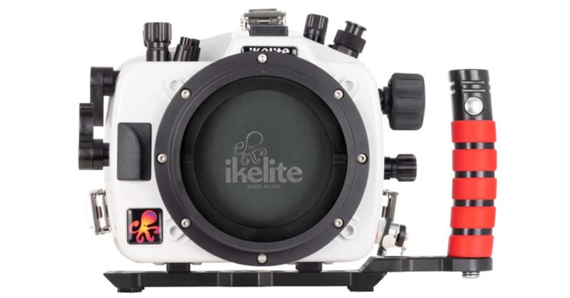 A white Ikelite camera housing with black accents, featuring a red and black striped handle on the right. The housing has several control knobs and a large circular lens area, with an octopus logo in the bottom left corner.
