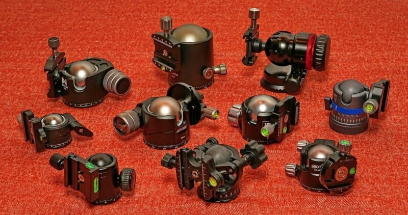 A collection of various tripod ball heads arranged on a red textured surface. Each ball head is black with different designs, knobs, and adjustment features, showcasing a variety of models and types commonly used in photography.