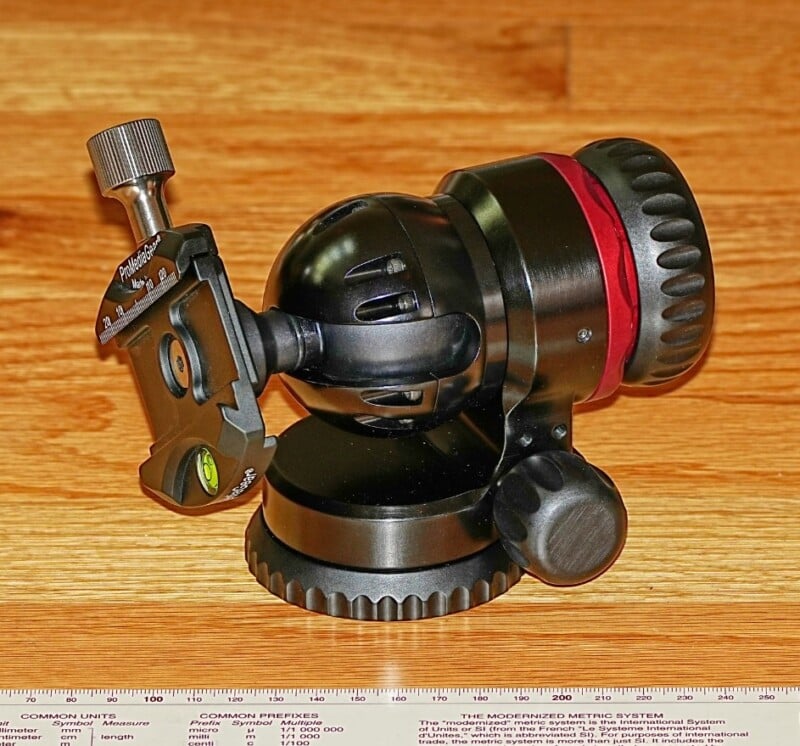 A close-up photo of a black and red camera tripod head with adjustment knobs and a leveling bubble, placed on a wooden surface. A ruler with metric and imperial units is visible at the bottom.